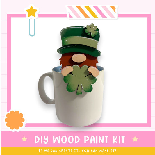 A mug with a small wooden gnome figure wearing a green hat and holding a green clover, set against a pink and white background.