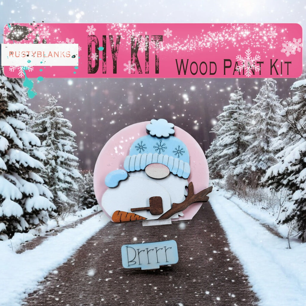 A DIY wood paint kit for creating a snowman, featuring a pink snowman figure and a snowman-themed sign. The background shows a snowy landscape with pine trees.