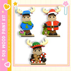 three wooden figurines of moose wearing different outfits, including a blue sweater, a Santa Claus outfit, and a green sweater.