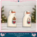 two snowman figurines with hearts in their pouches, set against a white wooden background with Christmas lights and pine trees in the background.