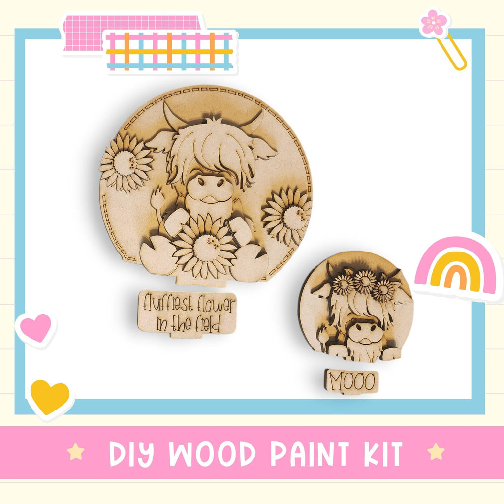 The image is a DIY wood paint kit featuring a wooden cutout of a cow holding a flower, with a smaller cutout of the same cow and a flower on it. The kit is presented in a pink and blue frame, and the text "DIY WOOD PAINT KIT" is displayed at the bottom.