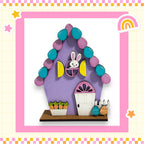 A colorful Easter-themed house made of various craft materials, including a purple and pink structure, a yellow door, and a white door. The house is decorated with Easter eggs and carrots, and there are small Easter eggs and carrots placed on the ground in front of the house.