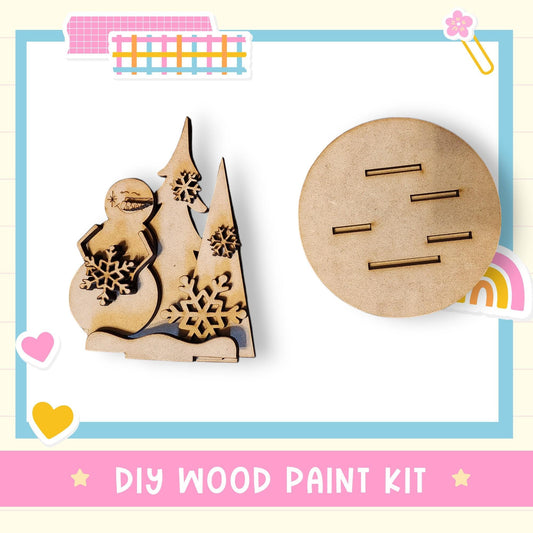 a DIY wood paint kit, including a snowman and a round wooden object with slats, all set against a white background with a pink border.