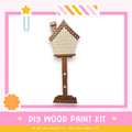 a DIY wood paint kit, with a wooden house-shaped object painted on a stand. The kit appears to be made of wood and is displayed against a pink and white background.