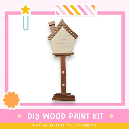 a DIY wood paint kit, with a wooden house-shaped object painted on a stand. The kit appears to be made of wood and is displayed against a pink and white background.