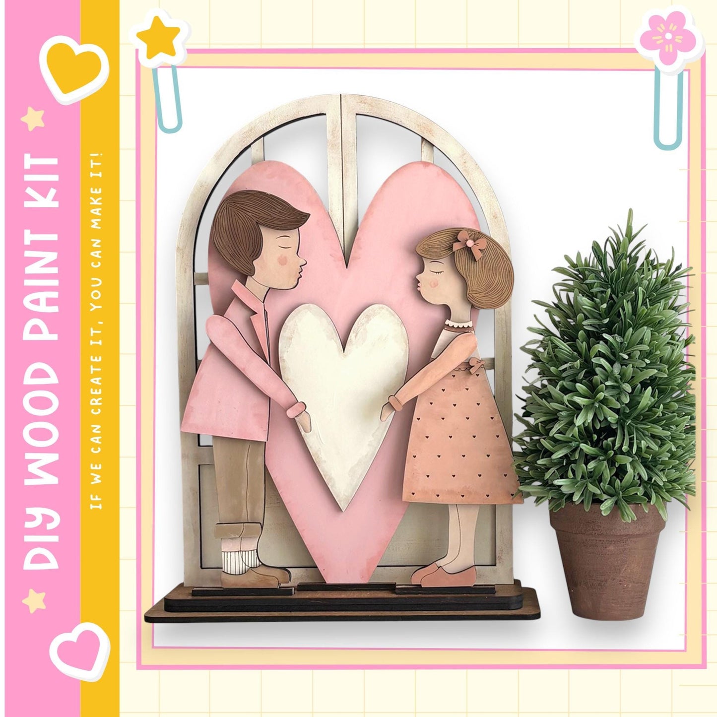 A wooden heart-shaped frame with two cartoon-like figures, one male and one female, standing inside it. The frame is placed on a shelf, and there is a potted plant to the right of the frame.