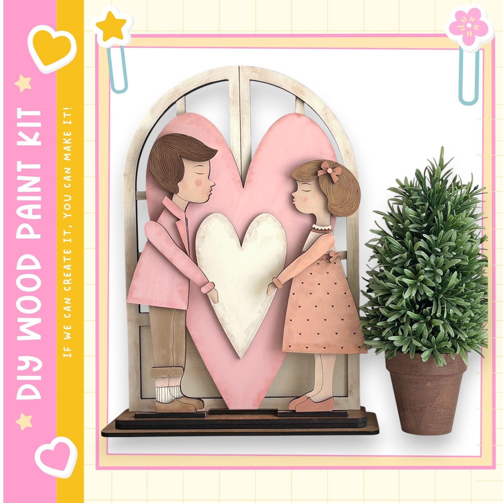 A wooden heart-shaped frame with two cartoon-like figures, one male and one female, standing inside it. The frame is placed on a shelf, and there is a potted plant to the right of the frame.