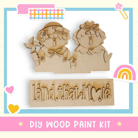 The image is a DIY wood paint kit featuring two wooden figures and a sign with the phrase "Land that I love".
