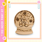 A wooden snow globe with a teddy bear design, surrounded by snowflakes, is displayed against a pink and yellow background.